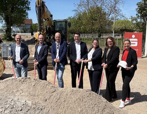 Die Gruppe steht vor einem Haufen mit Grund. Die Personen haben eine Spaten in der Hand. Im Hintergrund steht ein Aufsteller mit dem Sparkassenlogo und ein Bagger.