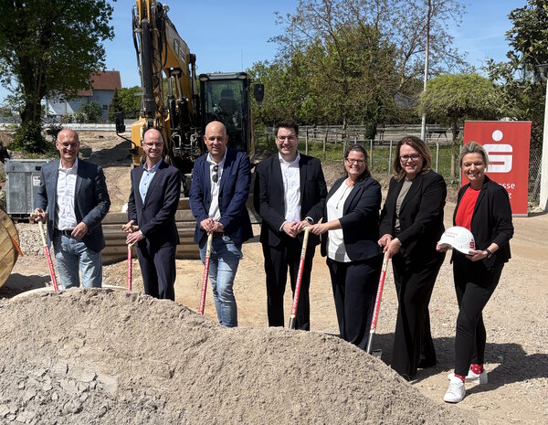 Bild vergr&ouml;&szlig;ern: Die Gruppe steht vor einem Haufen mit Grund. Die Personen haben eine Spaten in der Hand. Im Hintergrund steht ein Aufsteller mit dem Sparkassenlogo und ein Bagger.